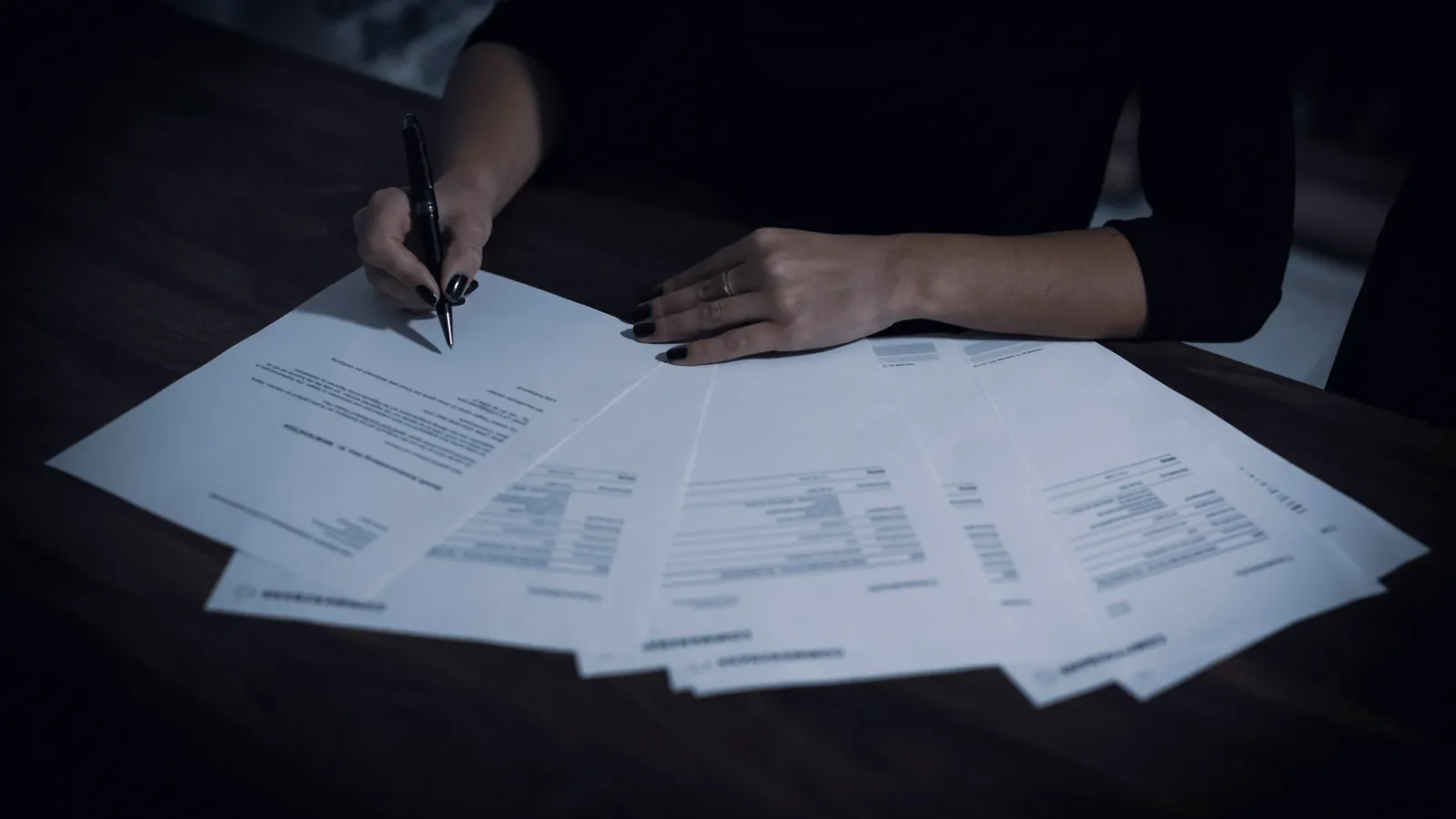 Mains qui signent des feuillets étalés sur une table sombre