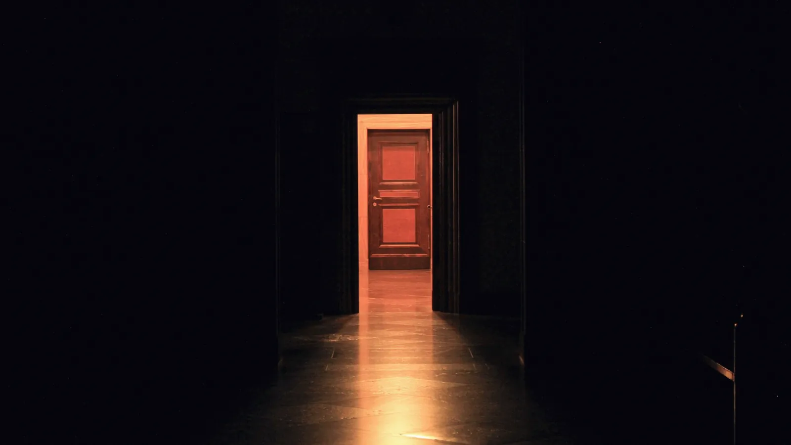Couloir sombre dont le fond s’ouvre sur une porte rouge entrouverte